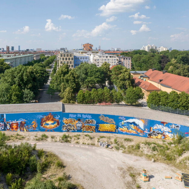 Drohnenaufnahme des Graffitis das sich längs über eine lange Wand zieht. Im HIntergrund sieht man eine Grünanlage und mehrere Wohnhäuser.