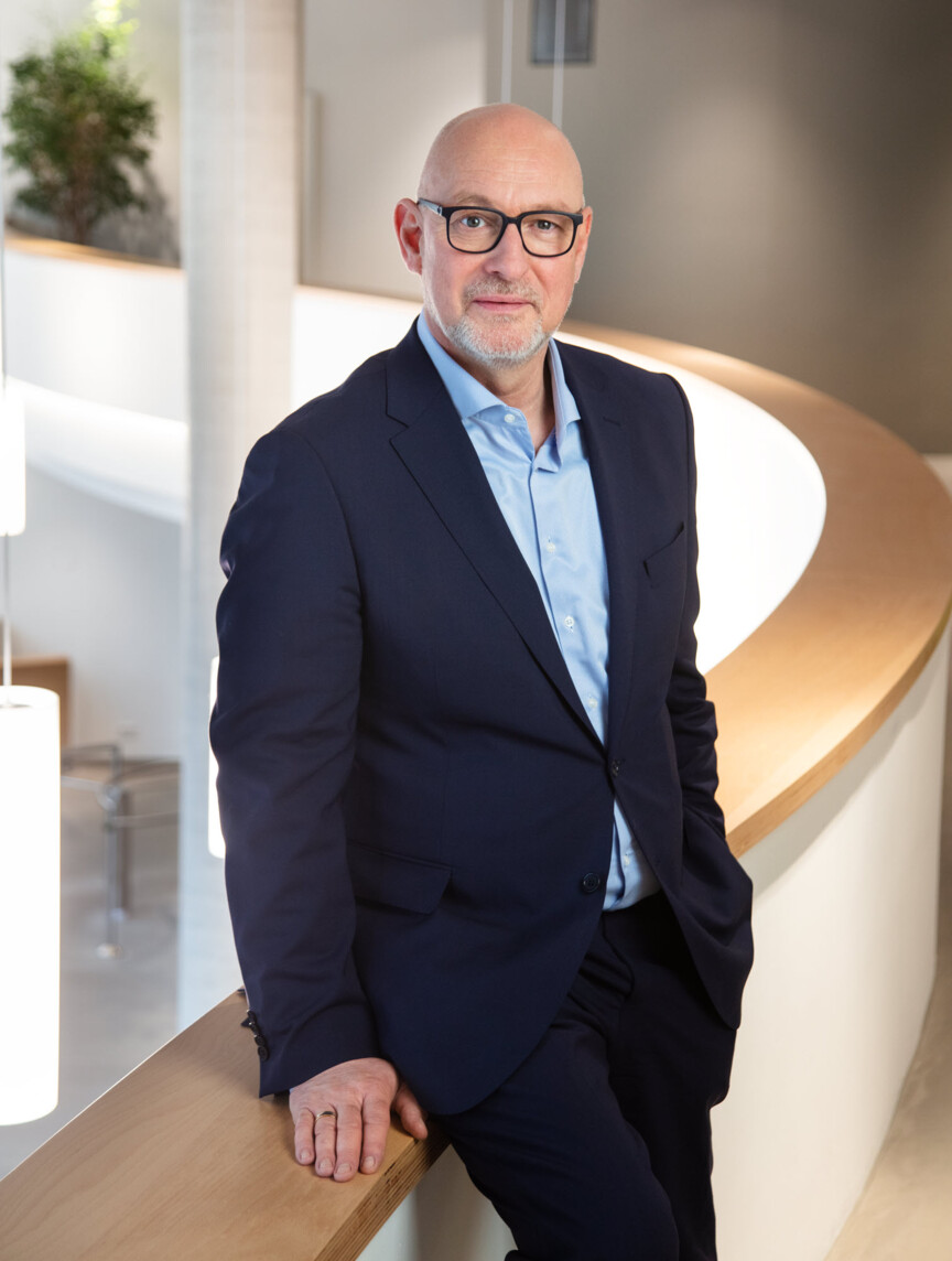 Portraitbild Jörg Franzen: Ein Mann mit Brille im dunkelblauen Anzug steht an eine Wand angelehnt.