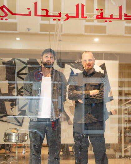 Zwei Männer stehen im Schaufenster eines Geschäfts- Im Hintergrund ist ein Künstleratelier und Installationen zu erkennen.