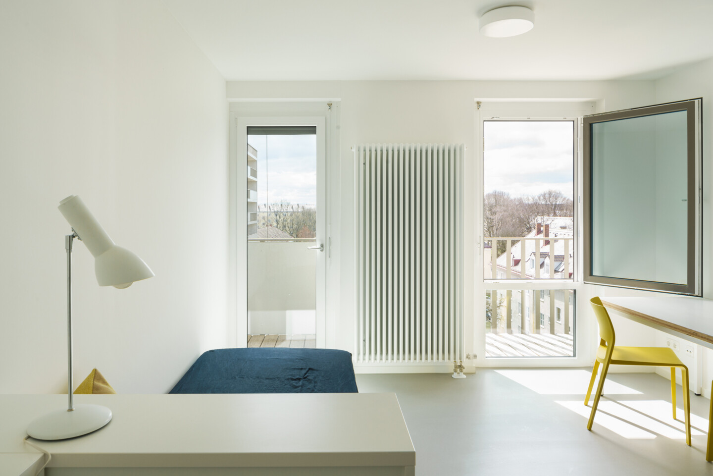 Blick in ein Einzelapartment. Rechts ist ein Tisch mit Stuhl zu sehen, links eine Kommode mit Lampe davor ein Bett. Am Ende des Raums sind zwei schmale bodentiefe Fenster und ein Heizkörper in der Mitte.