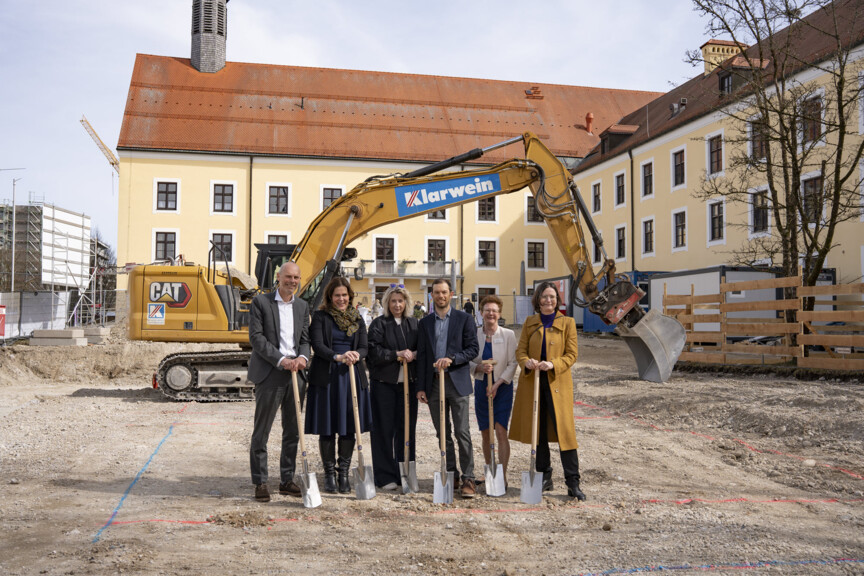 Gruppenfoto auf einer Baustelle vor einem Bagger. Die abgebildeten Personen stehen in der Bildunterschrift.