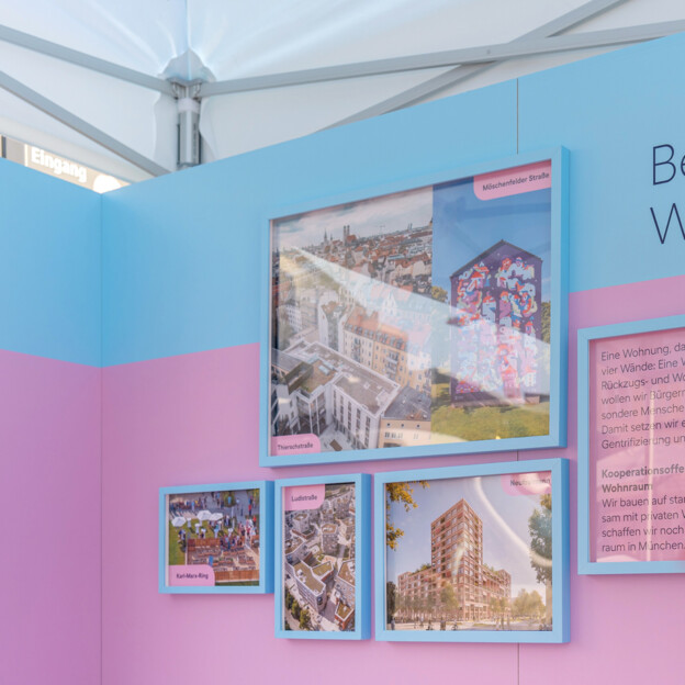 Blick auf einen Messestand auf dessen Wand steht "Bezahlbarer Wohnraum". Daneben hängen Fotografien von Neubauten und Quartieren. Im Vordergrund sieht man ein Windrad mit dem Münchner Wohnen Logo.