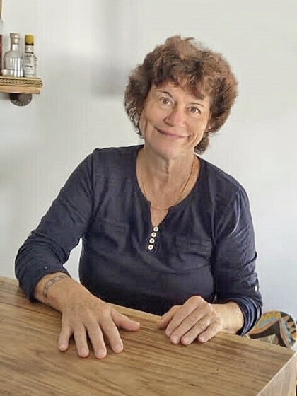 Portraitbild einer älteren Frau mit braunem kurzen Haar. Sie sitz am Tisch und lächelt in die Kamera.