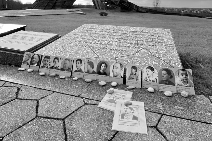 15 Fotografien von Münchner*innen, die nach Litauen verschleppt wurden, sind vor einer Gedenktafel aufgestellt. Davor liegt vor jedem Bild ein Stein.