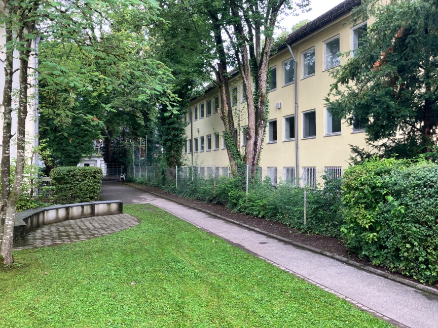 Blick auf ein dreistöckiges älteres Wohngebäude davor ein asphaltierter Weg und eine Rasenfläche.