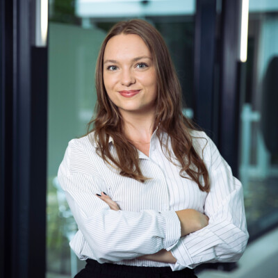 Portraitbild einer jungen Frau mit langen braunen Haaren. Sie steht in einem Büroumfeld und trägt eine gestreifte Bluse und eine schwarze Hose.