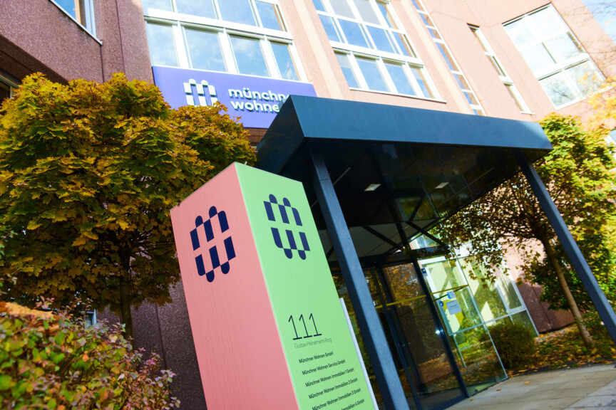 Blick auf den Eingang der Hauptverwaltung der Münchner Wohenn. Neben dem überdachten Eingang steht eine Stehle mit Münchner Wohnen Logo.