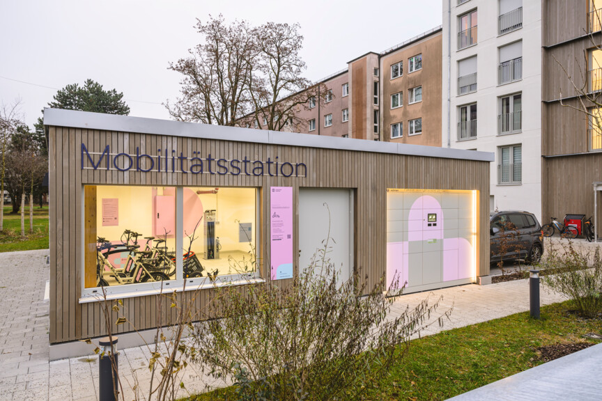 Blick auf eine Mobilitätsstation die zwischen zwei neuen Wohnhäuserin in Holzbau steht. Im großen fenster sind E-Bikes und weitere Ausleihgeräte zu sehen.