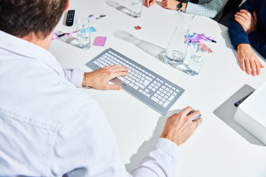 Blick über die Schulter eines Mannes auf einen Tisch auf dem eine Tastatur, eine Maus und Getränke stehen.