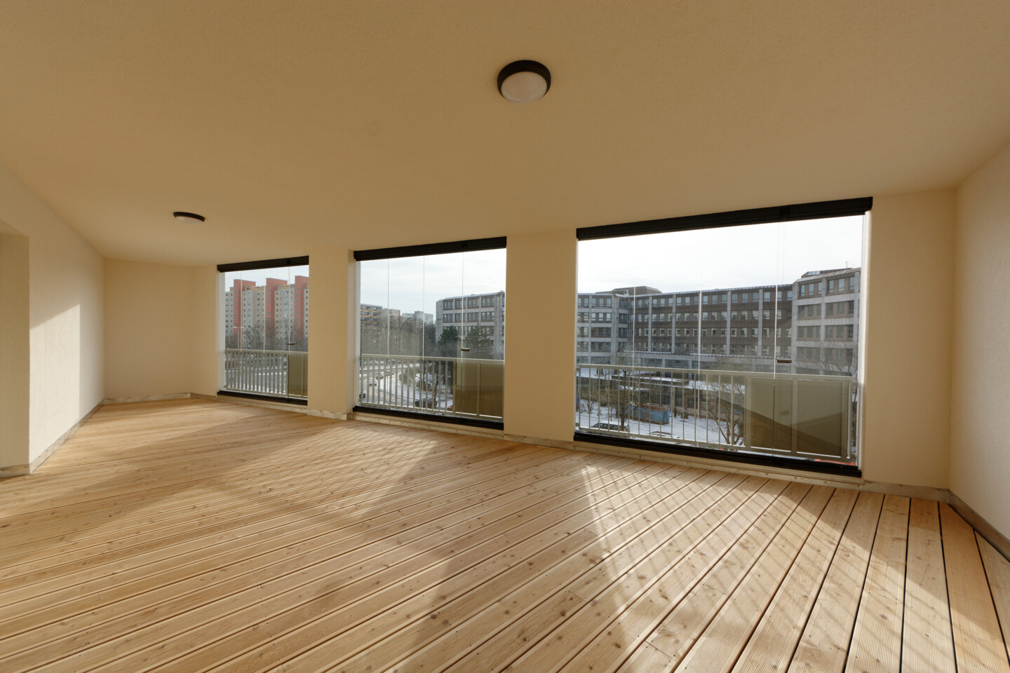 Blick in eine verglaste Loggia, die den Blick auf die Straße und das gegenüberliegende Gebäude freigibt. Am Boden sind helle Holzlatten verlegt.
