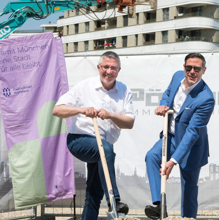 Zwei Männer setzen je einen Spaten in einen Geröllhaufen. Sie stehen auf einer Baustelle und sind umringt von Baggern und weiteren Baugeräten.