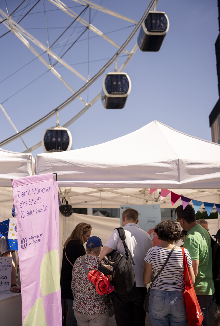 Blick auf den Stand der Münchner Wohnen mit einer Menschengruppe davor. Im Hintergrund sieht man die Gondeln eines Riesenrades.