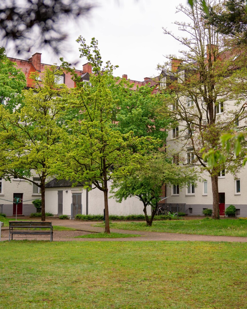 Blick in den Innenhof des Hohenzollernkarrees. Ein Gehweg zwischen vielen Bäumen und verschiedene Sitzgelegenheiten sin dzu sehen. Dahinter das mehrstöckige Wohngebäude.