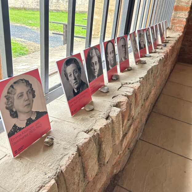 Mehrere Fotografien von Menschen stehen nebeneinander auf einer Mauer an ein Fenster angelegt. Vor jedem Bild liegt ein kleiner Stein.