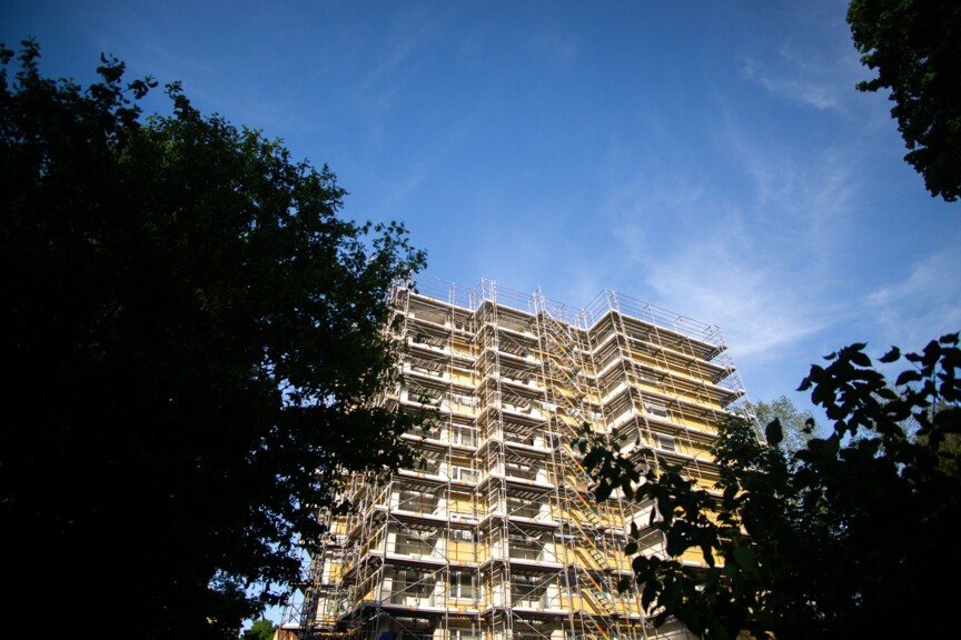 Blick von unten nach oben auf ein Hochhaus, das von Gerüsten umgeben ist. Links und rechts ist das Bild von Bäumen eingerahmt.