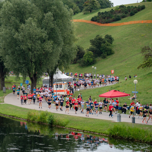 Viele Menschen joggen auf einem Weg entlang eines kleinen Sees. Am Streckenrand stehen weitere Menschen und jubeln ihnen zu.