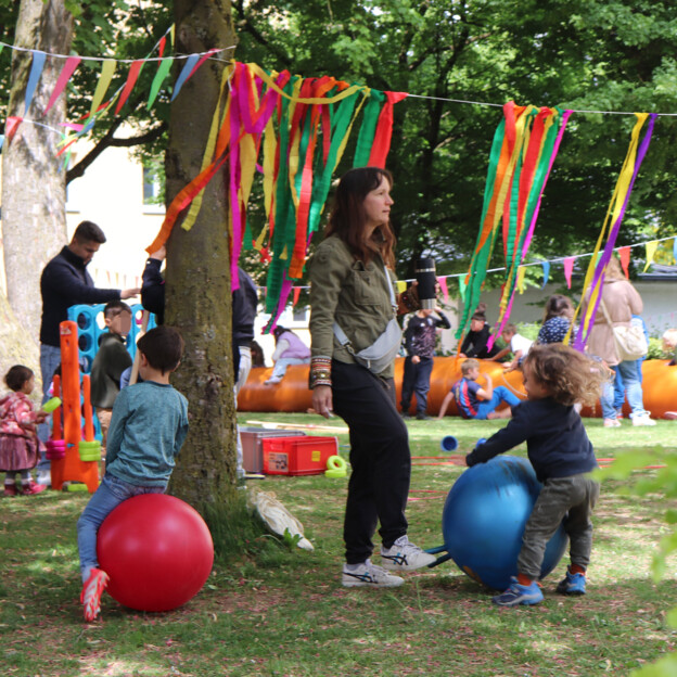 Blick auf eine Wiese auf der viele Kinder mit verschiedenen Spielsachen spielen. Dazwischen sind auch immer wieder Eltern.