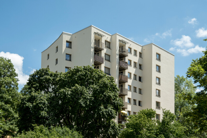 Ansicht eines hellen, mehrstöckigen Punkthauses von unten, mit übereinanderliegenden Balkonen an der Fassade, teils mit Sonnenschirm und Möbeln. Das Gebäude ragt vor blauem Himmel auf und wird im unteren Bildbereich von dichten, grünen Baumkronen umrahmt.
