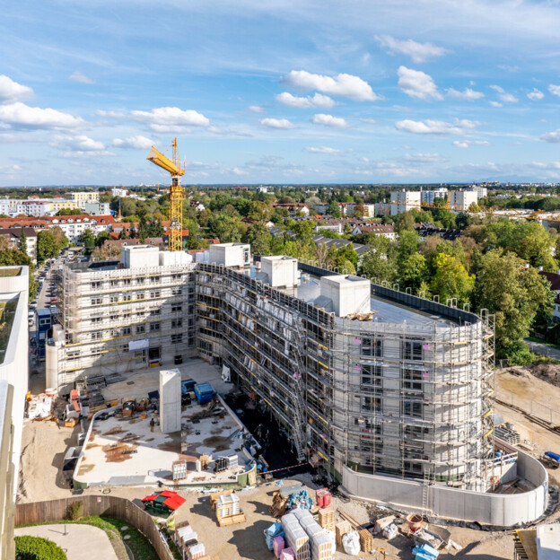 Drohnenaufnahme des L-förmigen Gebäudes um das noch Baugerüste stehen. IM Hintergrund viel Grün und weitere Wohngebäude.