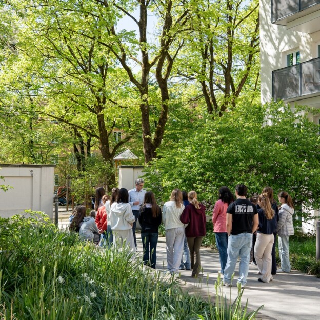 Eine Gruppe junger menschen hört einem Mann zu. Sie stehen im Innenhof einer Wohnanlage mit viel Grün.
