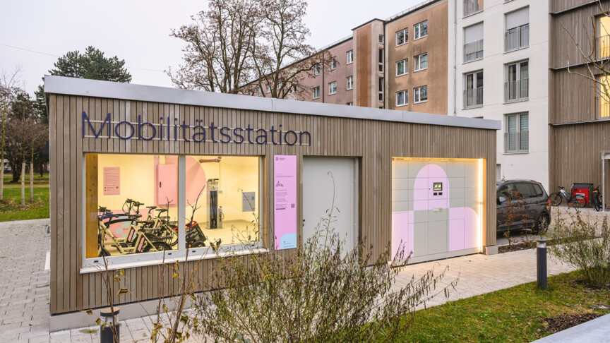 Blick auf eine Mobilitätsstation der Münchner Wohnen. Durch das große Fenster erkennt man mehrere E-Bikes und Lastenräder.