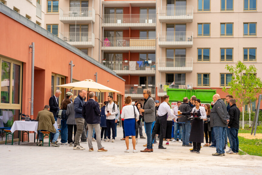 Blick auf die Wohnanlage. Im Vordergrund stehen Menschen in Gruppen zusammen und unterhalten sich.