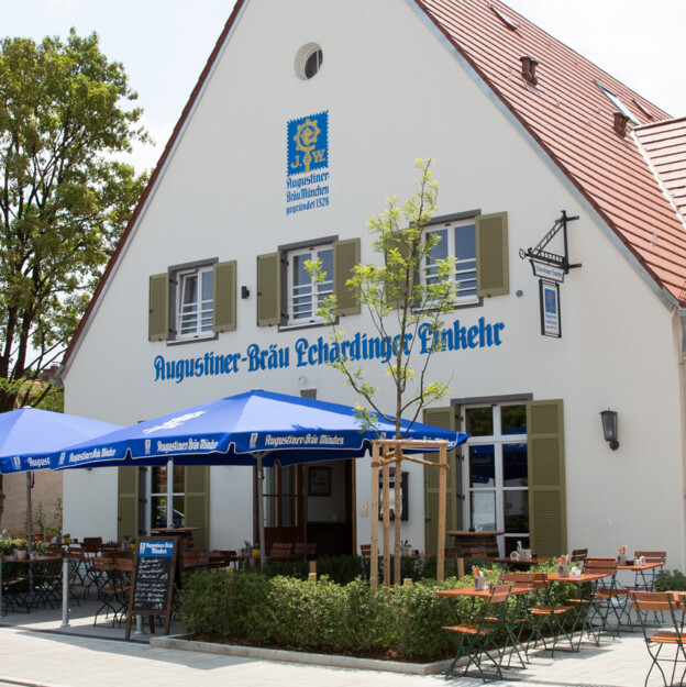 Blick auf ein Wirtshaus vor dessen Eingang Tische und Stühle unter Sonnenschirmen stehen.