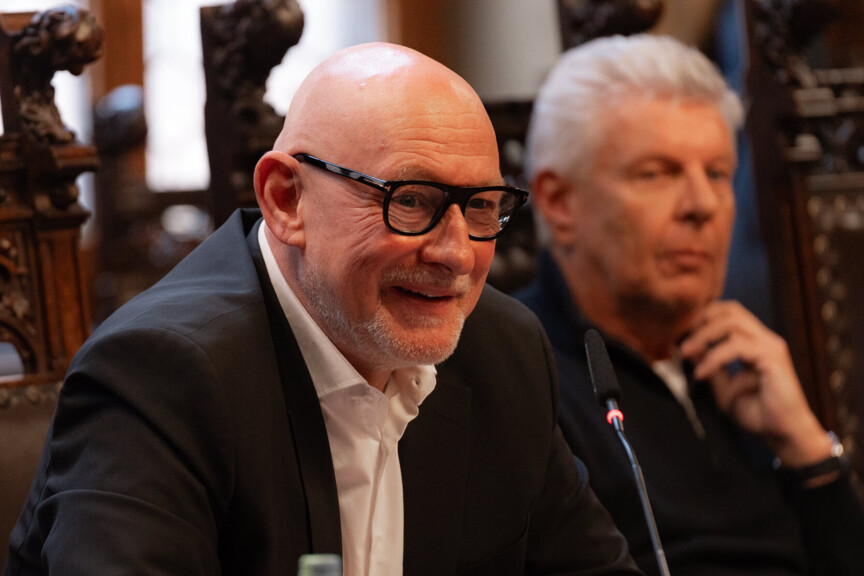 Zwei Männer sitzen nebeneinander. Einer von ihnen lehnt sich vor und spricht in ein Tischmikrofon.