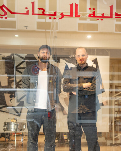 Zwei Männer stehen im Schaufenster eines Geschäfts- Im Hintergrund ist ein Künstleratelier und Installationen zu erkennen.