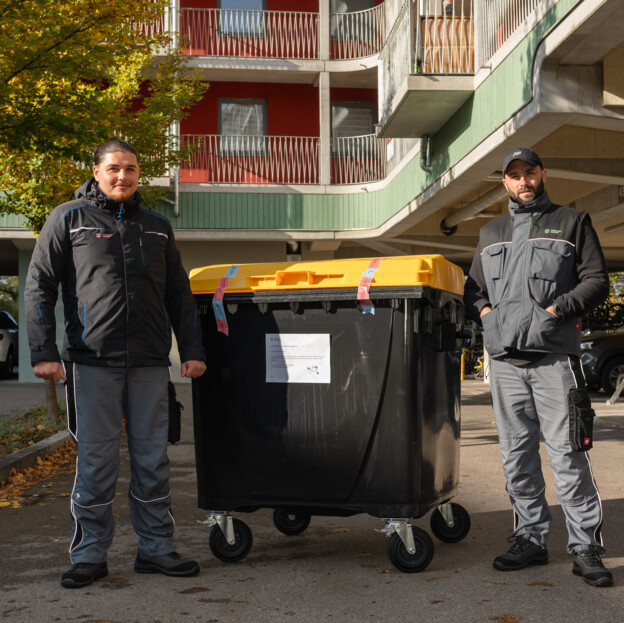 Zwei Männer in Arbeitskleidung der Münchner Wohnen stehen neben einer gelben Tonne.