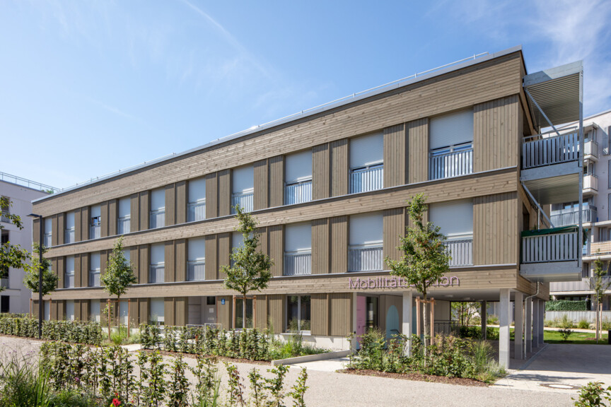 Blick auf ein modernes WOhngebäude mit Holzfassade und einer Mobilitätsstation im Erdgeschoss.