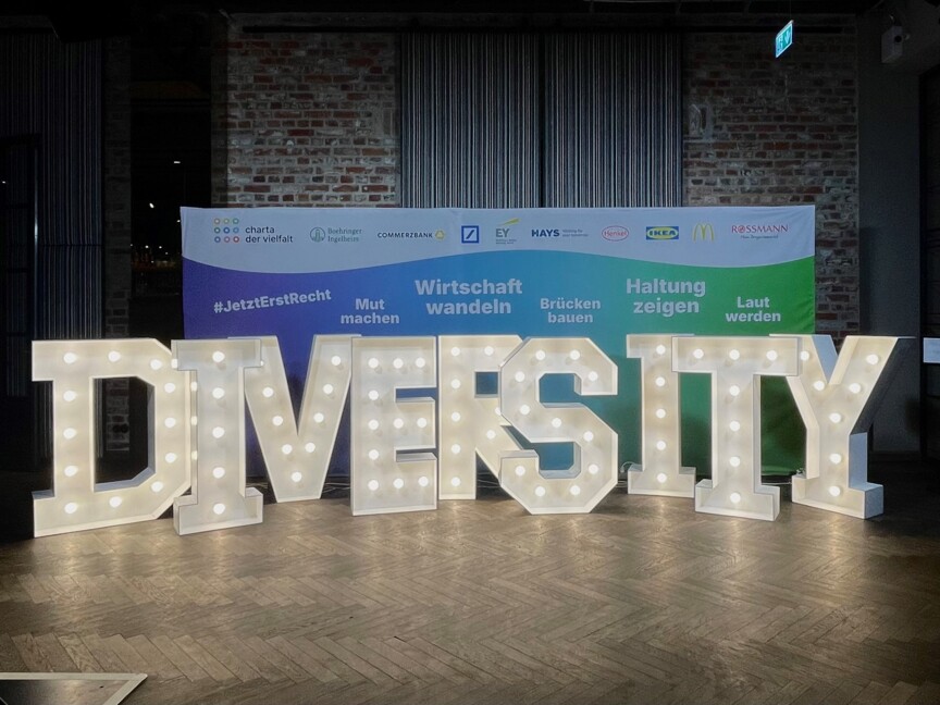 Große Leuchtbuchstaben stehen in einem Raum mit sehr hohen Decken. Sie bilden das Wort Diversity. Dahinter steht ein großes Banner mit den Logos verschiedener Firmen, die die Diversity-Konferenz gesponsert haben.