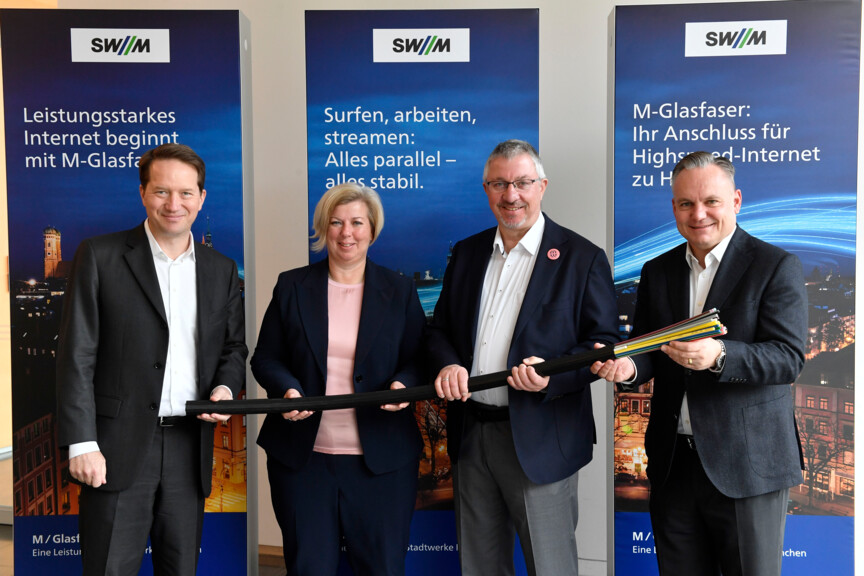 Drei Männer und eine Frau halten ein übergroßes Glasfaserkabel in der Hand. Hinter Ihnen stehen Werbebanner der Stadtwerke München.