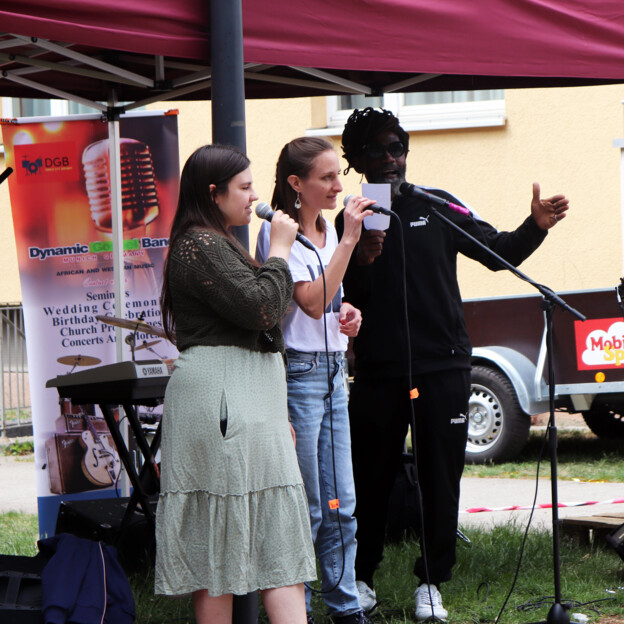 Blick auf drei Menschen die am Mikrofon singen. Dahinter steht ein Roll Up der Band "The Dynamic Band".