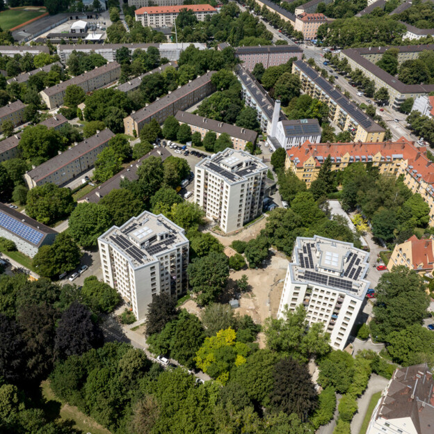 Luftaufnahme eines Wohngebiets mit mehreren hohen Punkthäusern mit Photovoltaikanlagen auf den Dächern, umgeben von dichtem Grün und Bäumen. Im Hintergrund erstreckt sich eine dichter bebaute Stadtlandschaft mit niedrigeren Wohnblocks, sowie Straßen und Parkplätzen.