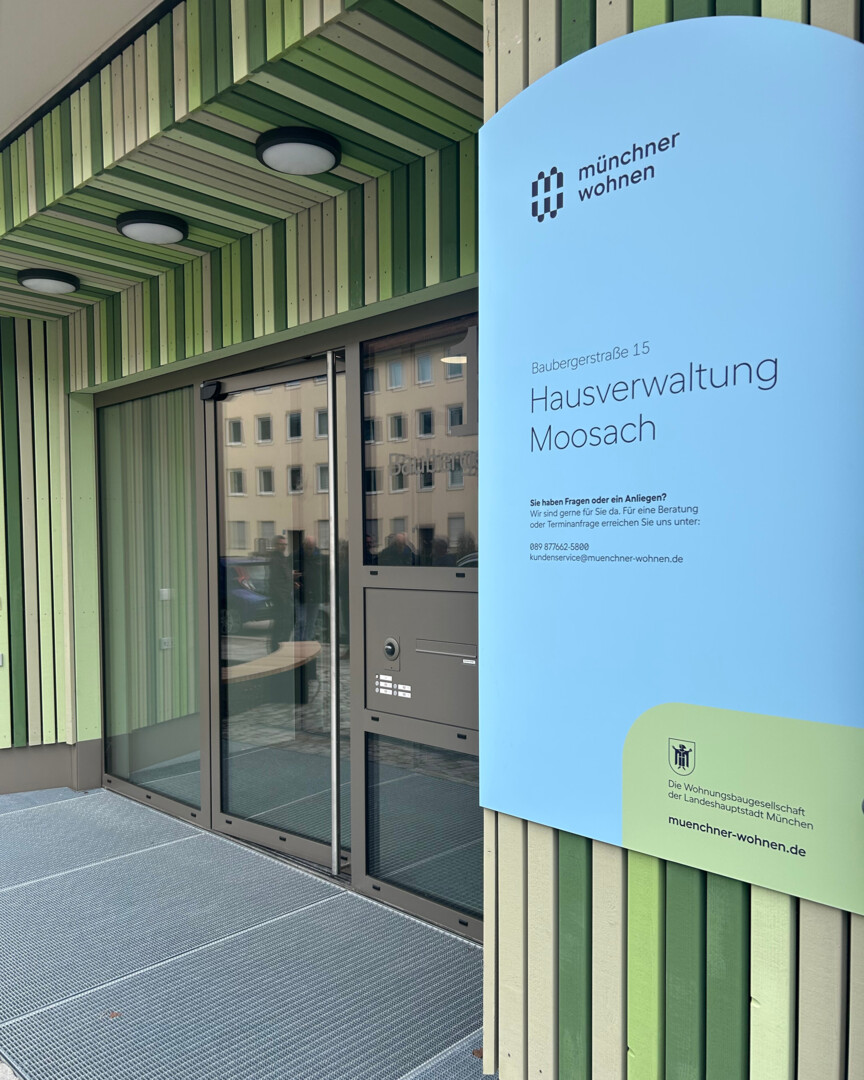 Blick auf den Eingang der Hausverwaltung Moosach. Rechts davon hängt ein Schild mit dem Logo, Namen und den Öffnungszeiten der Hausverwaltung.