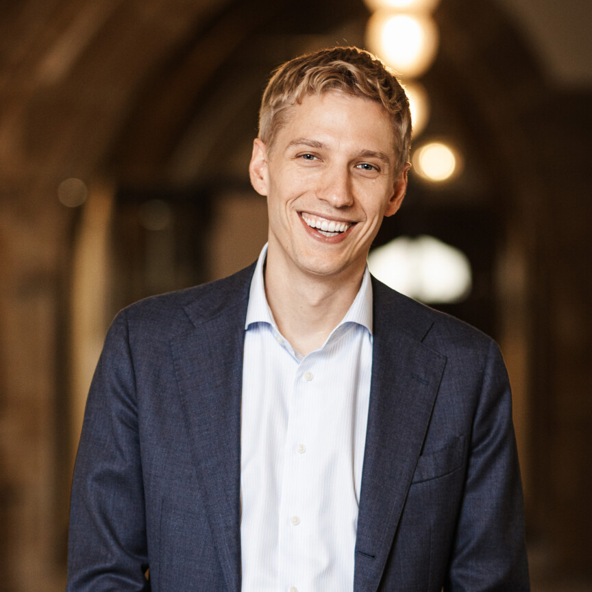 Ein junger blonder Mann im Anzug lächelt in die Kamera.