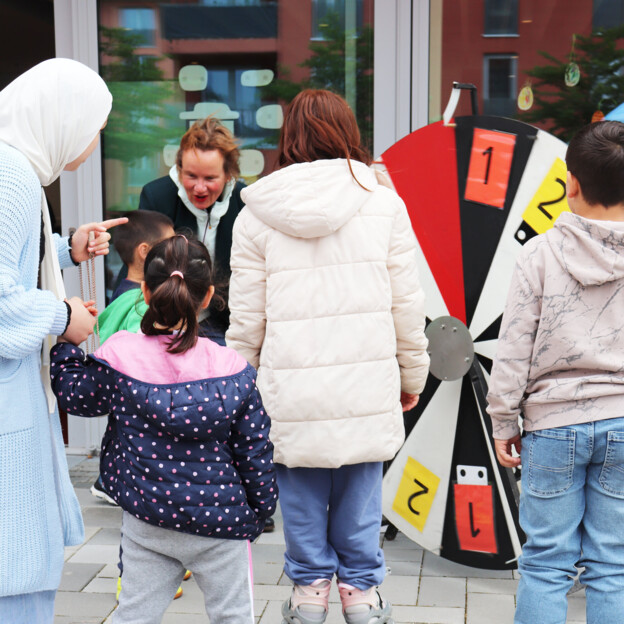Familien mit Kindern stehen vor einem Glücksrad. Eine Mitarbeiterin animiert die Kinder mitzumachen.