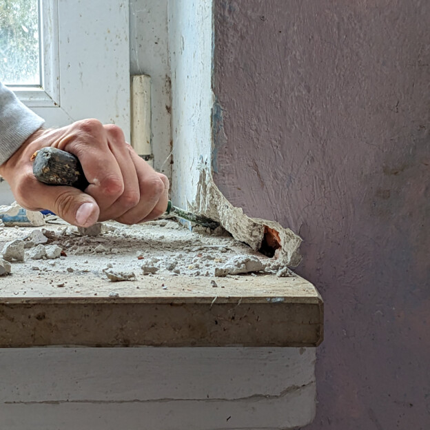 Blick auf ein Fensterbrett. Man sieht eine Hand die das Mauerwerk um das steinerne Fensterbrett zu entfernen und die Steinplatte zu lösen.