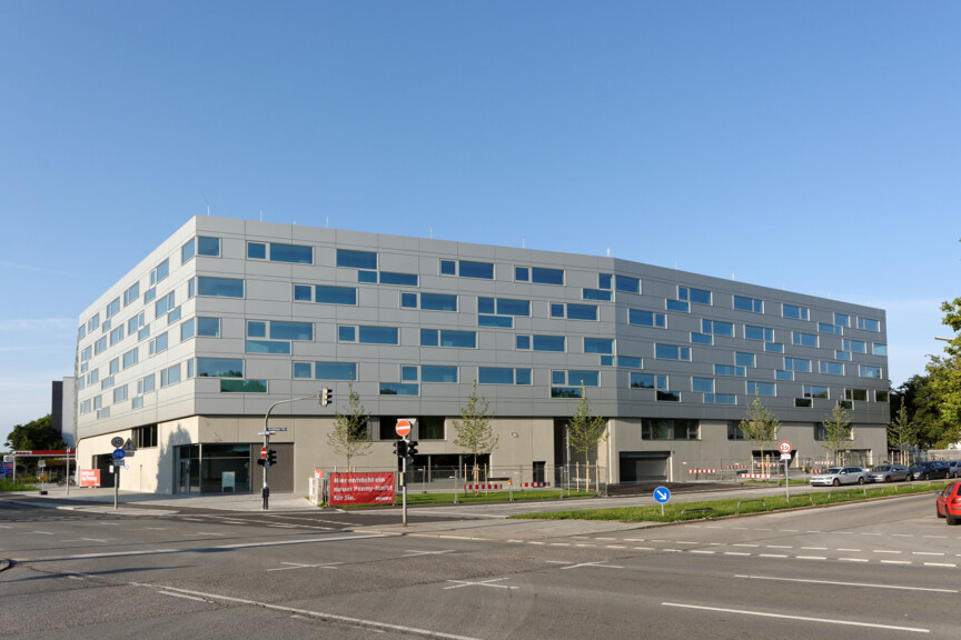 Blick auf das Wohnhaus mit den Azubiapartments. Ein modernes Haus das über Eck verläuft und eine dunkle Fassade mit schmalen Fenstern hat.