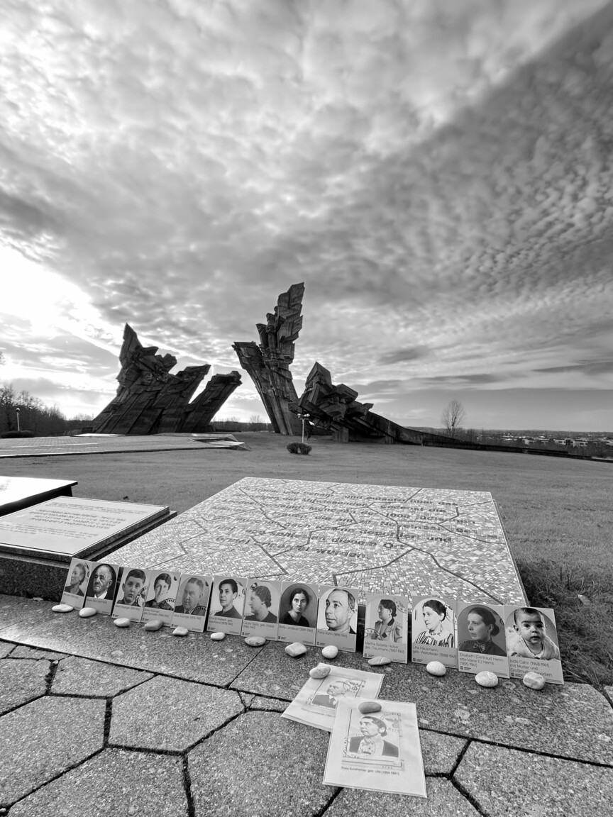 15 Fotografien von Münchner*innen, die nach Litauen verschleppt wurden, sind vor einer Gedenktafel aufgestellt. Davor liegt vor jedem Bild ein Stein.