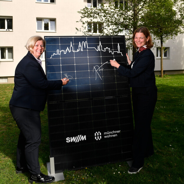 Zwei Frauen stehen im Innenhof einer Wohnanlage. Zwischen ihnen halten sie ein Solarpanel und unterschreiben darauf.