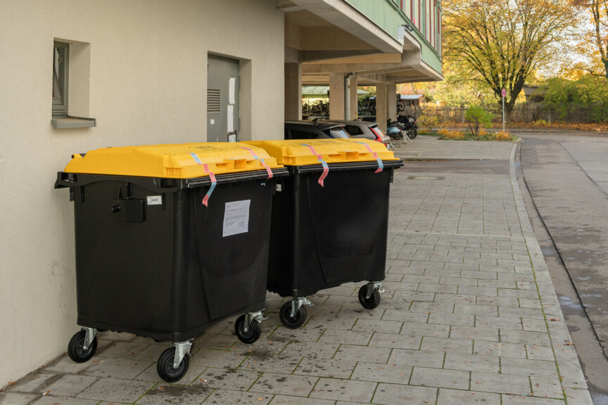 Zwei große Mülltonnen mit gelbem Deckel und mit Klebeband verschlossen stehen auf dem Gehweg vor einem Mehrfamilienhaus.