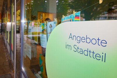 Blick auf das Schaufenster des Nachbarschaftstreffs. Auf einer hellgrün hinterlegten Fläche steht "Angebote im Stadtteil".
