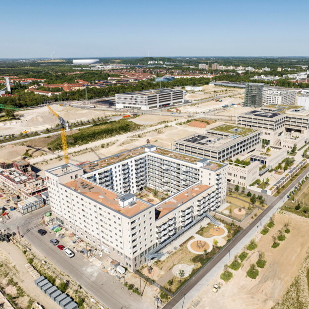 Luftaufnahme des fertiggestellten Wohnkomplexes aus größerer Höhe mit Blick auf das umliegende Neubaugebiet. Im Hintergrund sind weitere Gebäude, Baustellen sowie am Horizont die Allianz Arena zu erkennen.