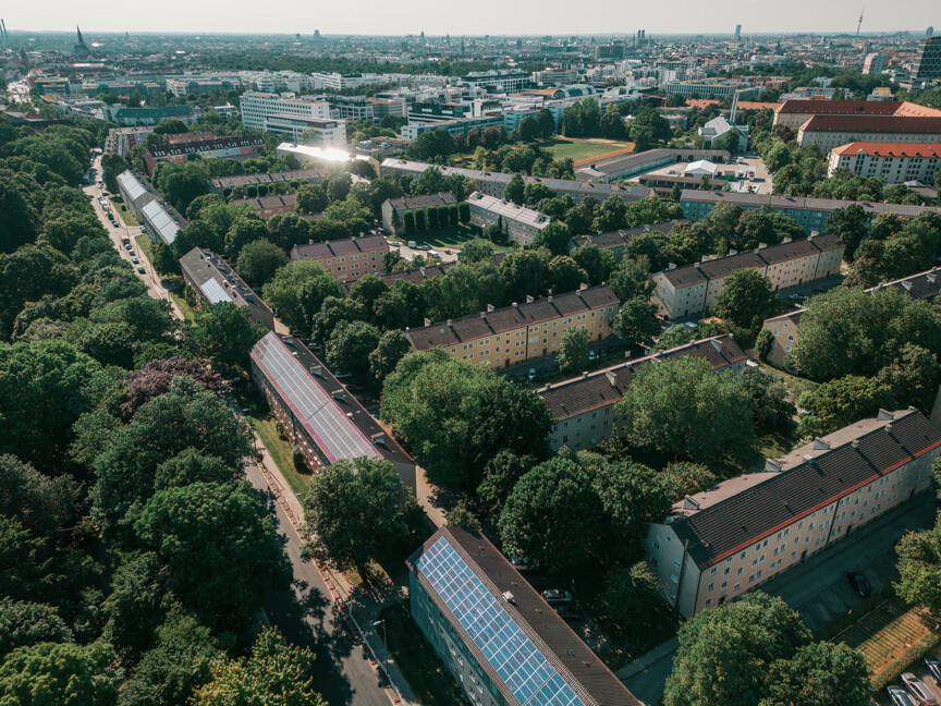 Luftbildaufnahme des Klimaquartier Ramersdorf. Auf dem Tortenstückförmigen Gebiet steht ein älterer Gebäudebestand. Der bereits modernisierte Part hat Photovoltaikflächen auf dem Dach. Zwischen den Häusern ist viel gewachsener Baumbestand.