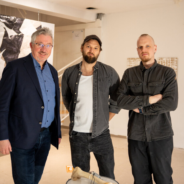 Drei Männer stehen in einem Künstleratelier und blicken in die Kamera.